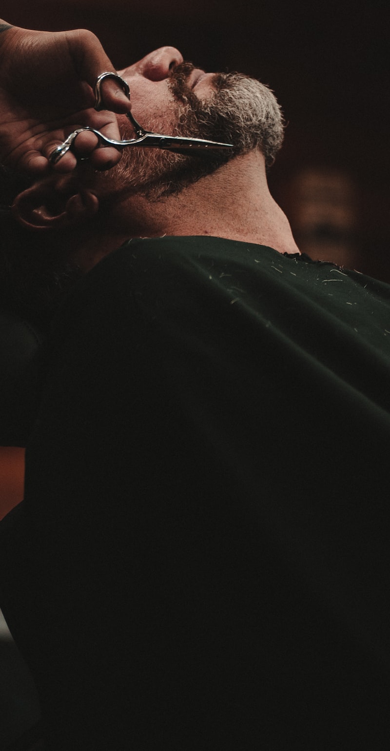 A barber trimming a reclined customer's beard with scissors